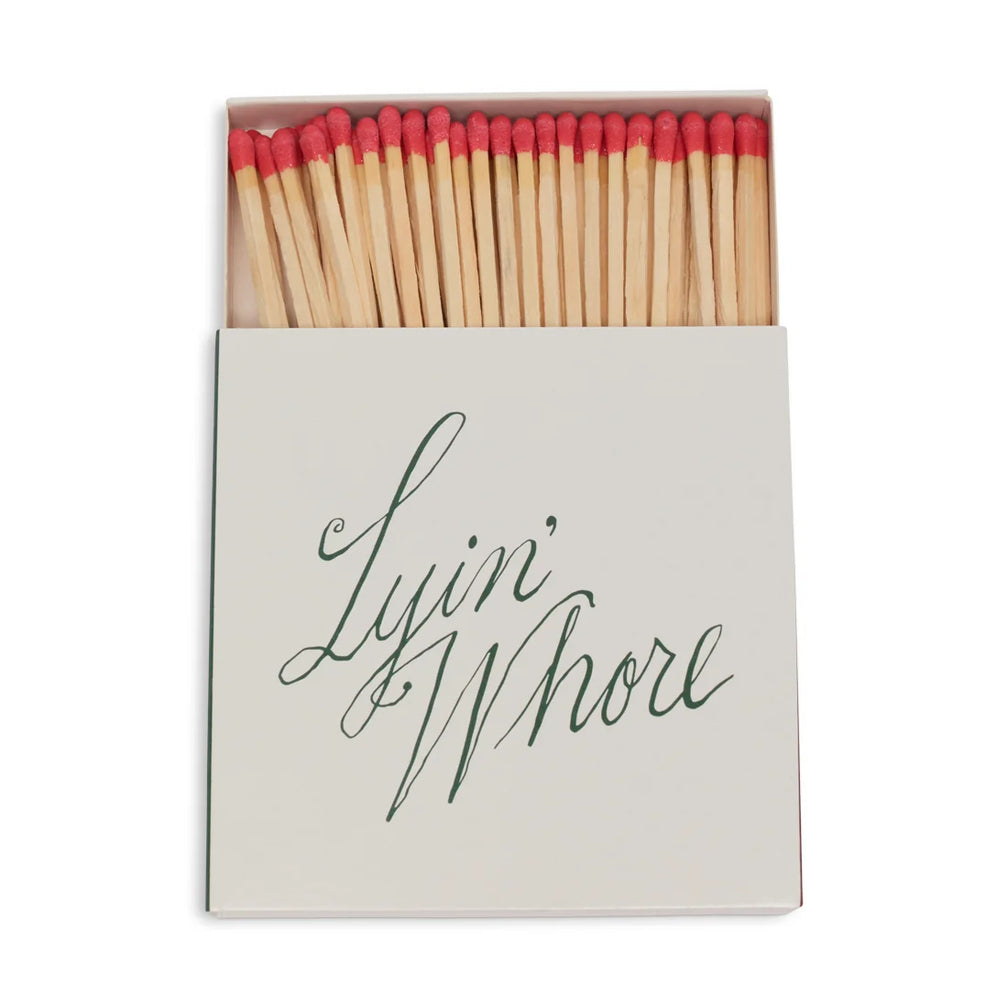 Square box of long red-tipped matches, open, white cover with Lyin' Whore in green cursive