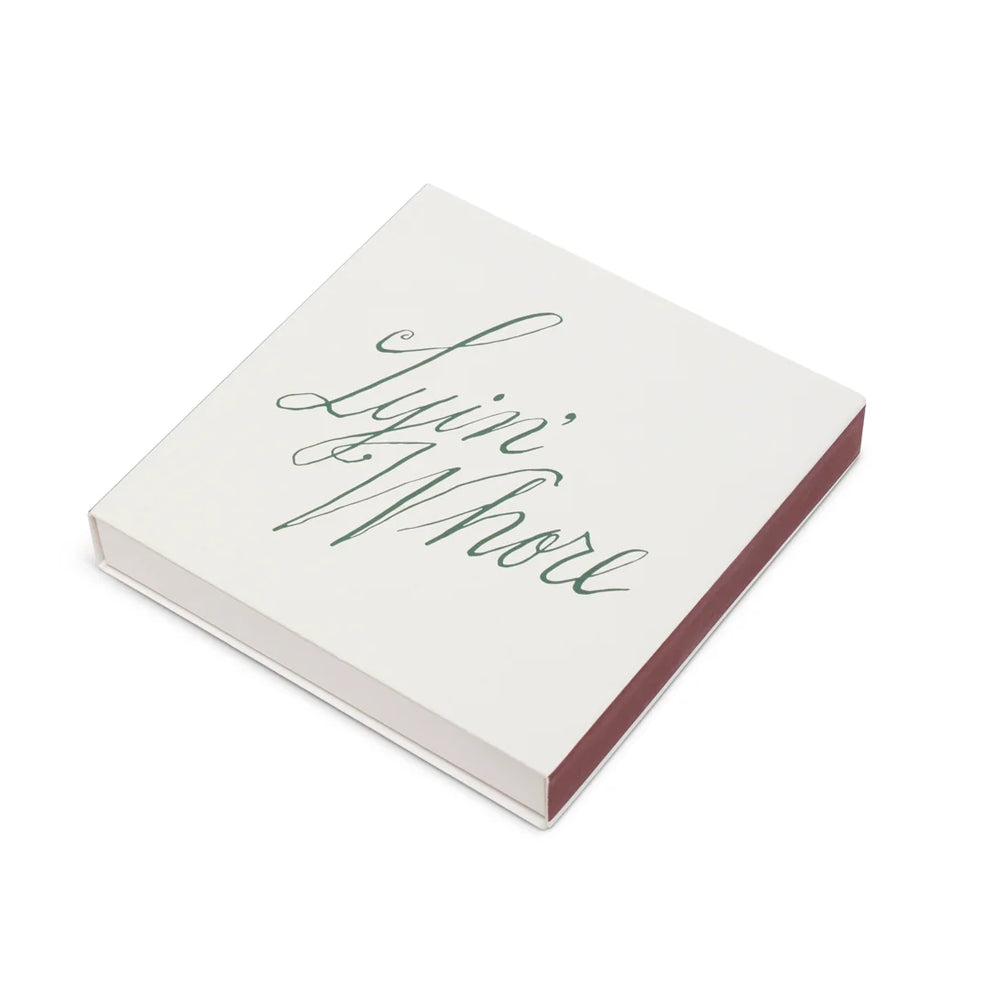 White square matchbox with Lyin' Whore green cursive script, angled to show the striker side