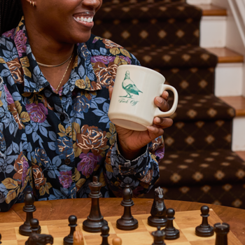 Person holding a mug with a green dragon design, sitting at a chessboard.