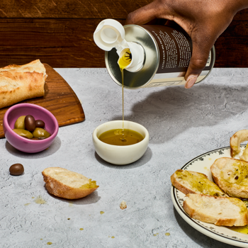 Person pouring olive oil from a can into a small bowl on a table with bread and olives.