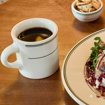 Mug of coffee on a wooden table with a plate of food and a bowl of snacks.