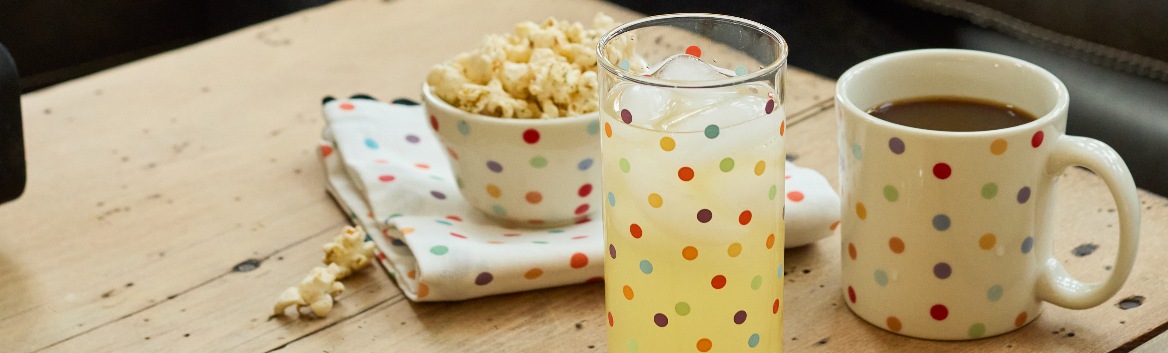 Polka dot mug, glass, and bowl on a wooden table with popcorn.