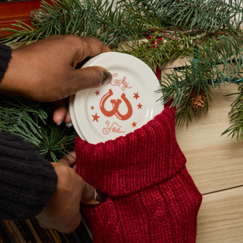 Hand placing a small white plate with red design into a red knitted stocking against a Christmas tree background.