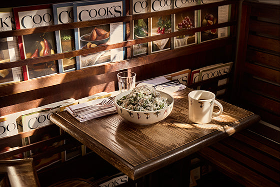 Wooden table with a bowl of salad and a mug, set against a wall with 'Cook's' magazine display.