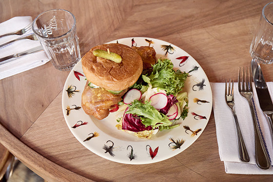 Plated dish with a sandwich and salad on a decorative plate with fly fishing-themed design, placed on a wooden table.