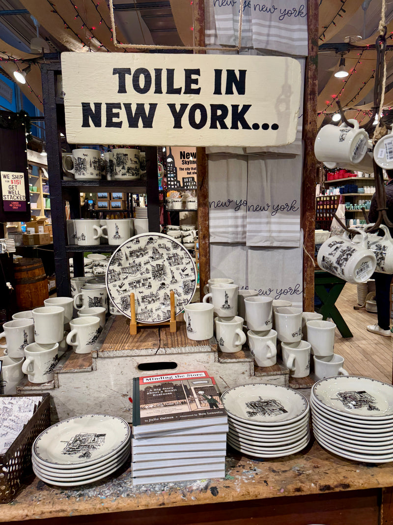 Display of 'Toile in New York' products including plates, mugs, and towels in a store setting.