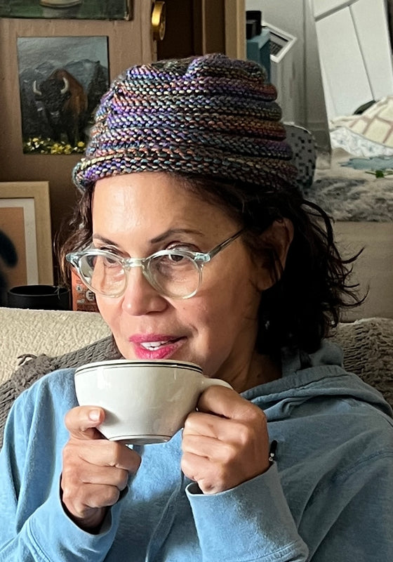 Person wearing a colorful knit hat and glasses, holding a mug in a cozy indoor setting.